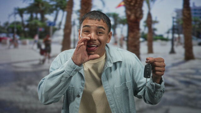 Man holding car keys, hand to mouth shouting and smiling on street with palm trees and promenade; excitement celebration pride.