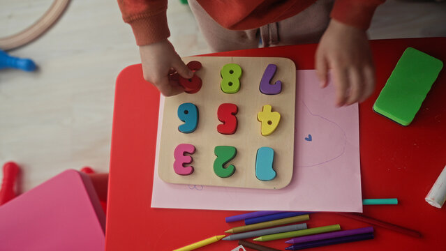 Boy using hands to place colorful wooden number puzzle pieces on red table inside building; playful early learning.