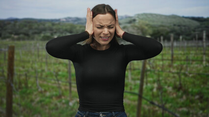 Obraz premium Woman in black top and jeans pressing hands to ears, eyes closed and mouth open in forest vineyard; emotional distress.