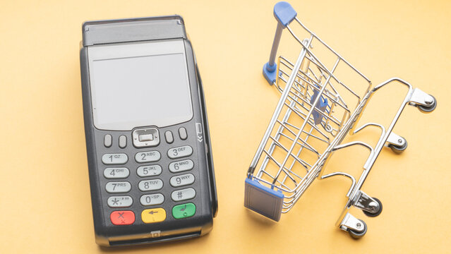 Black POS terminal with screen and buttons sits on yellow surface. Mini metal shopping cart stands nearby. No people present in image. Bright background emphasizes commercial elements clearly
