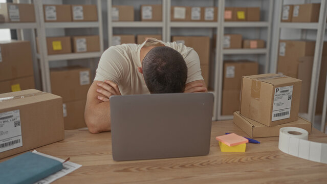 Man at desk surrounded by parcel boxes, sitting with hand on forehead and laptop in a building; frustration small business.
