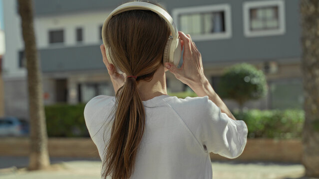 Young woman wearing headphones adjusts earcup while standing on a street near a building; serenity.