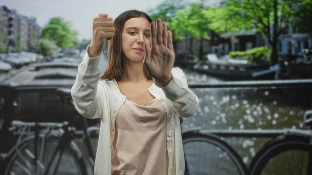 Woman with palm forward and clenched fist miming steering on a street by a canal and bicycle bridge; playful curiosity.