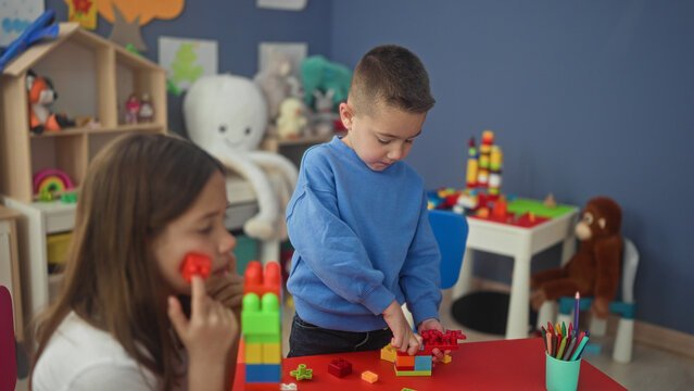 Boy stacking colorful plastic blocks with hands on a red table in a playroom inside a building while girl watches and crayons sit in a cup; quiet concentration.