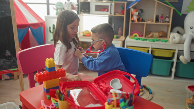 Girl and boy roleplay doctor with red stethoscope, boy listens to girl's chest and adjusts earpiece at a playroom; playful care.