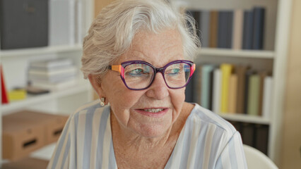 Woman wearing colorful glasses smiles and looks to the side in a building with bookshelves and a desk  serene reflection. © Krakenimages.com