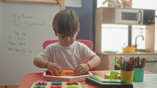 Boy using hands to sort a wooden shape sorter on a red table beside a whiteboard that reads virtual school in a home classroom; concentration learning play.