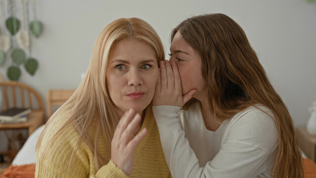 Blonde mother listens as her daughter whispers into her ear with hand covering mouth while seated on a bed in a bedroom; surprise intimacy trust.