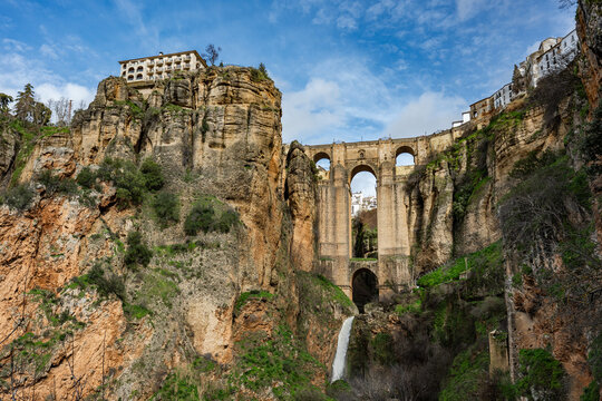 Wide angle view from the bottom of the gorge featuring the massive arches of Puente Nuevo bridge and a small waterfall in Ronda, Spain.