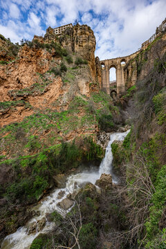 Wide angle view from the bottom of the gorge featuring the massive arches of Puente Nuevo bridge and a small waterfall in Ronda, Spain.