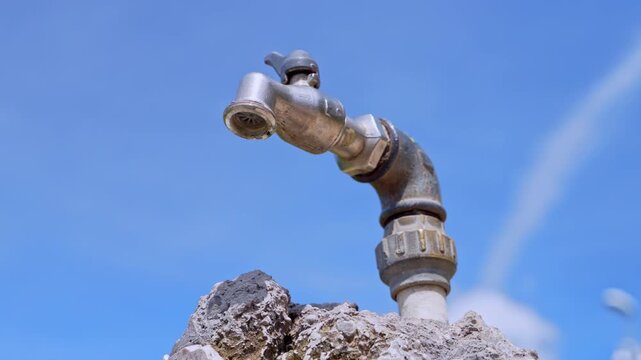 Old metal water tap on a stone against blue sky outdoors with no running water, single drop hanging, water scarcity, saving and ecology concept, drought awareness.