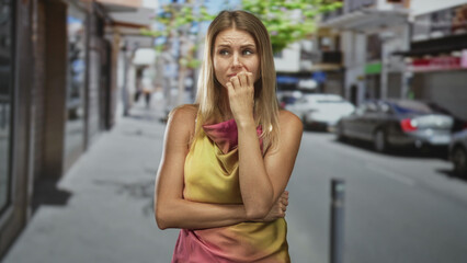 Fototapeta premium Woman biting her nails, hand to mouth and arms crossed on a street outside storefronts, wearing yellow and pink dress; anxiety isolation.