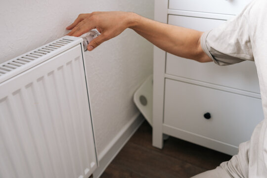Closeup of male hand adjusting thermostat on white radiator. Home heating control, energy saving and utility cost management concept. Person regulating room temperature for comfort.