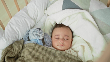 Naklejka premium Baby boy sleeping with closed eyes in a crib, face visible, swaddled under a green blanket beside a plush rabbit inside a building; serene rest.
