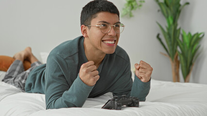 Man lying on bed holding a joystick with clenched fists, smiling and wearing glasses beside a gamepad on a white duvet in a studio  excitement. © Krakenimages.com