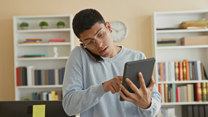 Young man on phone at ear while holding tablet, finger tapping screen in study with bookshelves and clock visible  focused multitasking. © Krakenimages.com