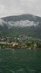 Fototapeta premium Lakeside village with waterfront buildings, rippling emerald lake water and low clouds clinging to the mountain slopes; serenity.
