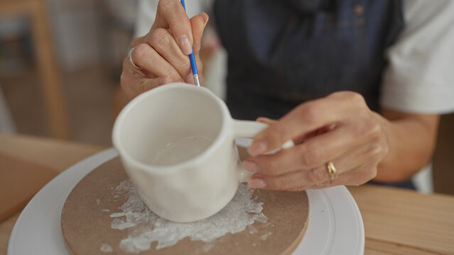 Woman painting ceramic mug with brush, hand holding handle in studio; concentration craftsmanship skill.