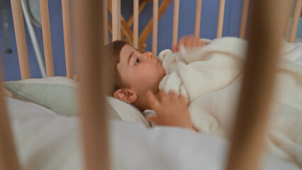 Boy lying in pale wooden crib clutching soft fleece blanket, hand near mouth, gaze sideways toward room window in building  cozy calm. © Krakenimages.com