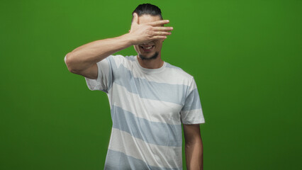 Fototapeta premium Man with hand covering eyes and smiling, striped tshirt in a studio green screen setting; playful concealment.
