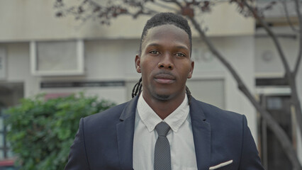 Fototapeta premium Man in navy suit with tie looks at camera on street near urban building and shrubs, standing upright with composed expression and direct gaze; confidence ambition professionalism.