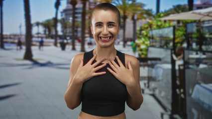 Fototapeta premium Woman short hair smiling, hands on chest and raised fists, wearing black crop top with bare midriff on street lined with palm trees; joy confidence.