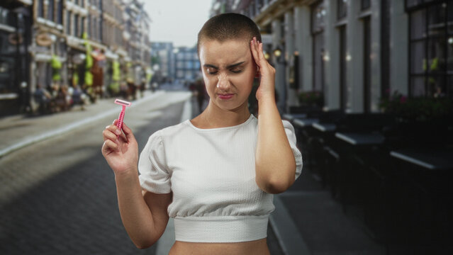 Woman with shaved head holding pink razor, hand to temple for headache while standing on street; self care discomfort.