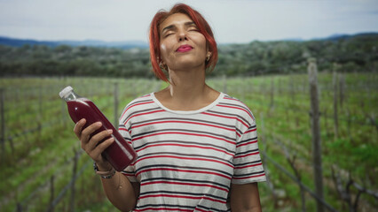 Obraz premium Woman holding a bottle of red juice with eyes closed in a vineyard field outdoors, striped shirt and bracelets visible; serenity.