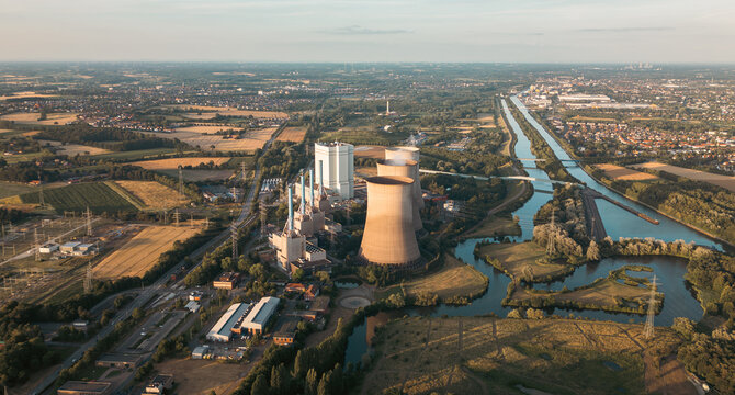 Industrial power plant with cooling towers surrounded by green fields and a canal under a clear blue sky, captured at sunset &mdash; symbol of modern energy and landscape harmony.