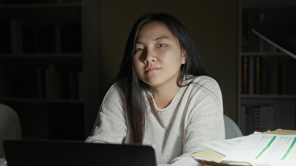 Naklejka premium Woman studying on laptop with paperwork and folders at desk in building, leaning forward toward the screen with a tired expression; late night fatigue.