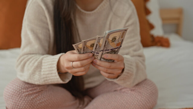 Woman counting dollars with hands while sitting crosslegged on a bedroom bed in pajamas; savings comfort.
