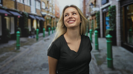Fototapeta premium Woman smiling with bare shoulders on a cobbled street, hands behind back, wearing black v neck tee; joyful casual confidence.