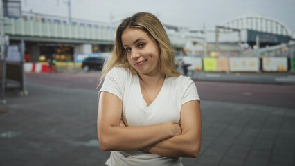 Fototapeta premium Woman with blonde hair and arms crossed on city street wearing white t shirt looking skeptical and unimpressed, subtle smirk; defiance doubt.