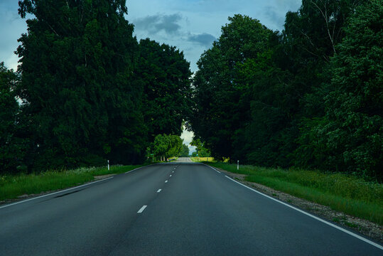 Empty rural highway stretching through farmland beneath dramatic storm clouds with forests and open countryside on the horizon.