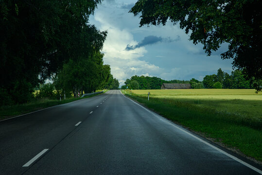 Empty rural highway stretching through farmland beneath dramatic storm clouds with forests and open countryside on the horizon.