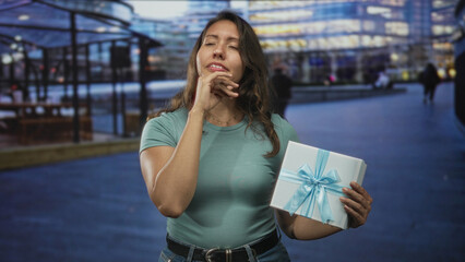Fototapeta premium Young latina woman holding a gift box with hand on forehead on street in front of building, eyes closed showing discomfort for headache; disappointment fatigue headache.
