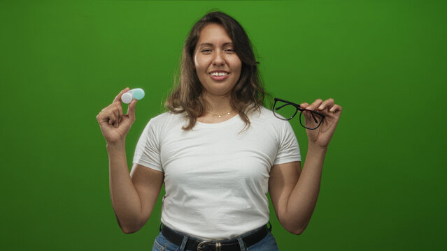Woman holding a contact lens case and eyeglasses while squinting her eyes in a green studio setting showing indecision and comparison; playful choice.