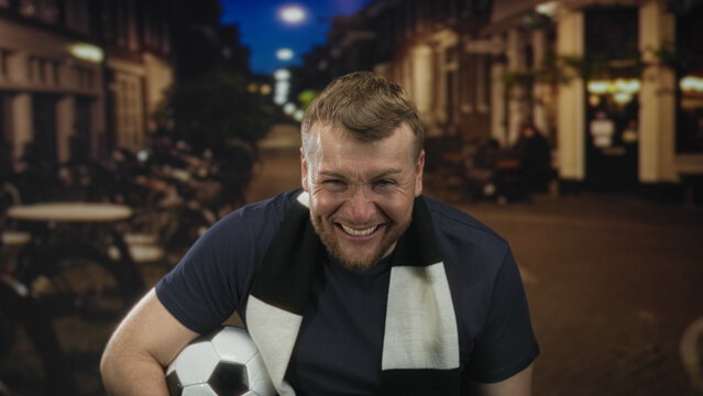Man holds soccer ball and laughs, hand covering face, wearing black and white scarf in a city street at night; team pride joy.