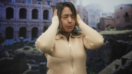 Fototapeta premium Hispanic woman covering ears with hands in front of ancient building ruins; sensory overload annoyance.