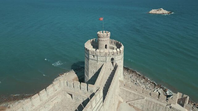 Aerial drone view of a historical coastal castle with a waving Turkish flag by the blue sea.