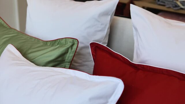 Layered decorative pillows fill cozy indoor scene, with white, green, and red cushions on upholstered seating beside softly blurred shelf of folded textiles, warm ambient light, smooth fabric texture