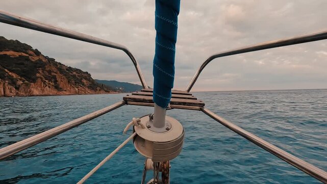 V&iacute;deo grabado desde la proa de un velero en navegaci&oacute;n, con la barandilla y el m&aacute;stil en primer plano, el mar abierto al frente y una costa rocosa con acantilados a un lado. La escena transmite traves