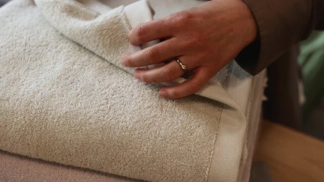 Close view shows hands holding and gently adjusting folded cream towel or thick textile beside brown clothing in soft indoor light, with plush texture, warm neutral palette, calm domestic mood, and