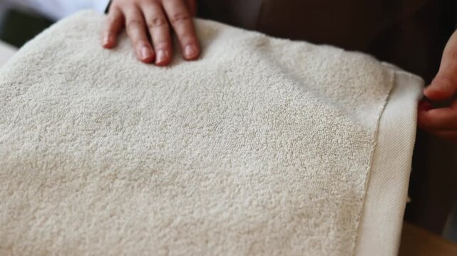 Two adult hands hold folded beige terry towel in close indoor view, showing careful lifting, positioning, and gentle fabric handling with warm light, clean domestic laundry mood, and visible texture