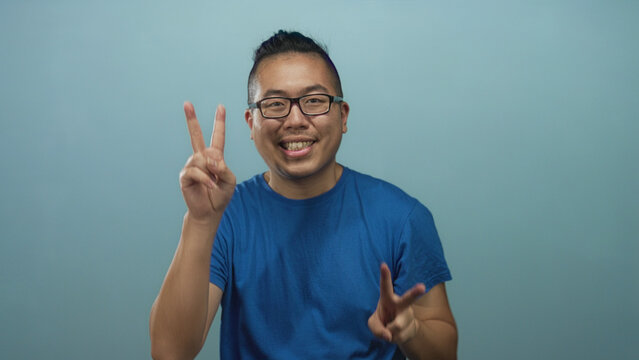 Young chinese man wearing blue t shirt and glasses smiling while flashing two peace signs with hands in studio against light blue wall; joy playful energy.