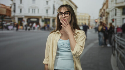 Fototapeta premium Woman wearing tortoiseshell glasses touching chin on city street, light blue top and beige overshirt among blurred buildings and a crowd; pensive reflection.