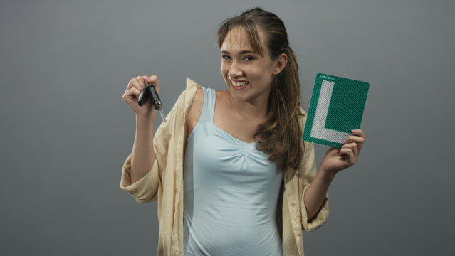 Woman smiling while holding car keys and green learner plate in grey studio, showing relaxed pose and casual shirt; pride new driver.