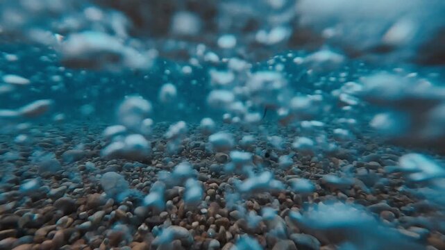 V&iacute;deo art&iacute;stico grabado desde la orilla, con olas rompiendo sobre la playa y la c&aacute;mara sumergi&eacute;ndose bajo el agua. Se aprecian burbujas, movimiento turbulento y piedras del fondo, creando una escena a
