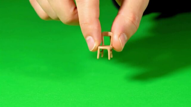 Close-up of a person's hand picking up and moving a tiny miniature chair toy on a green screen background