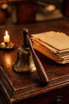A vintage brass school bell with a wooden handle rests on a rustic dark wooden desk beside a stack of old envelopes and a burning candle.